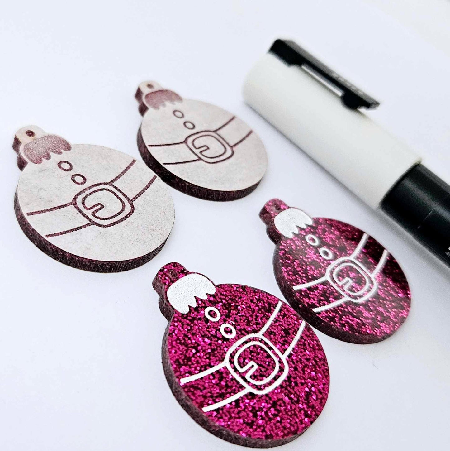 One pair of hot pink glitter Santa bauble dangle earring blanks paint filled white, alongside one unpainted pair with protective paper attached, pictured with a Posca paint pen