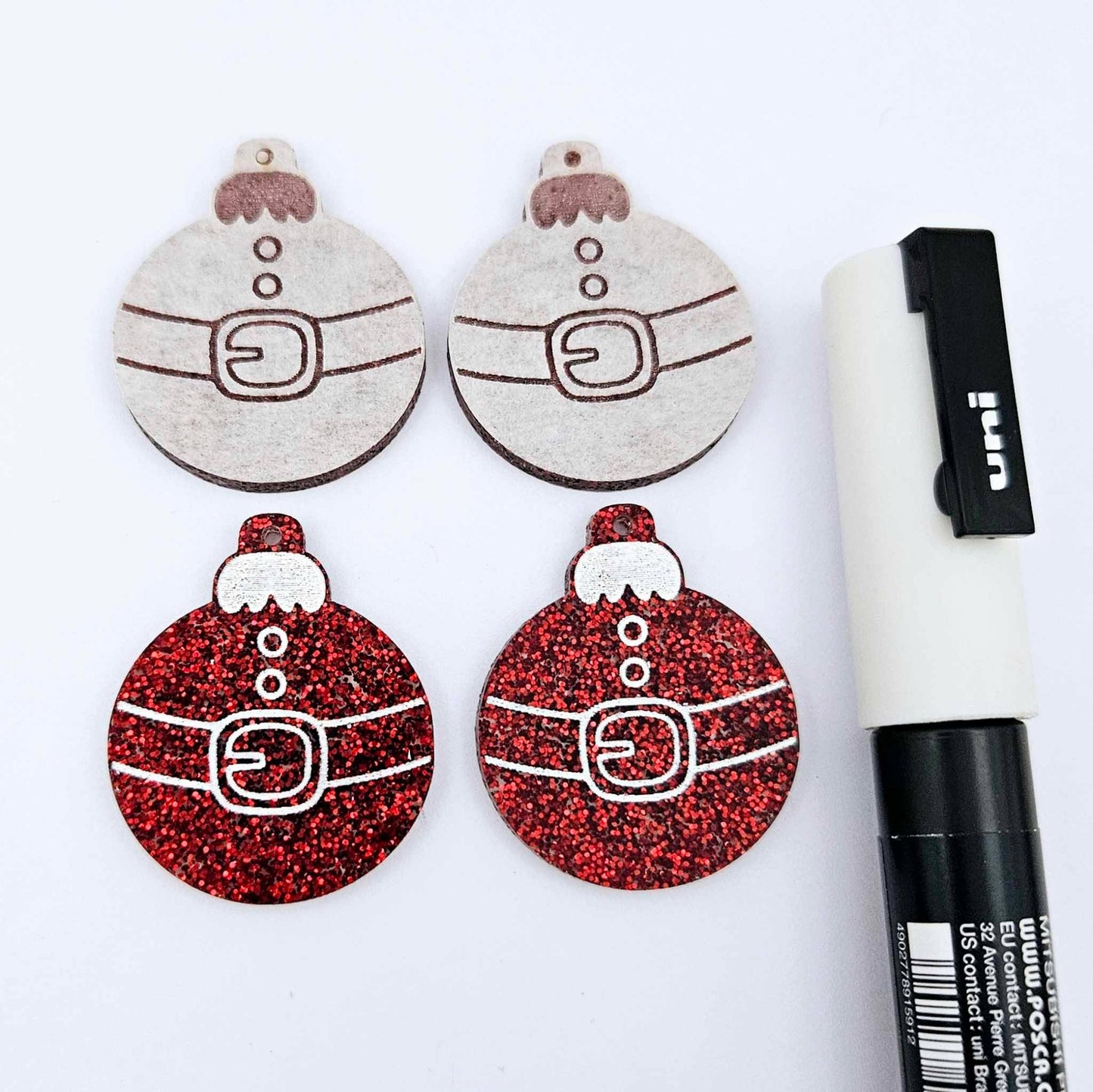 One pair of red glitter Santa bauble dangle blanks paint filled white, alongside one unpainted pair with protective paper attached, pictured with Posca paint pens