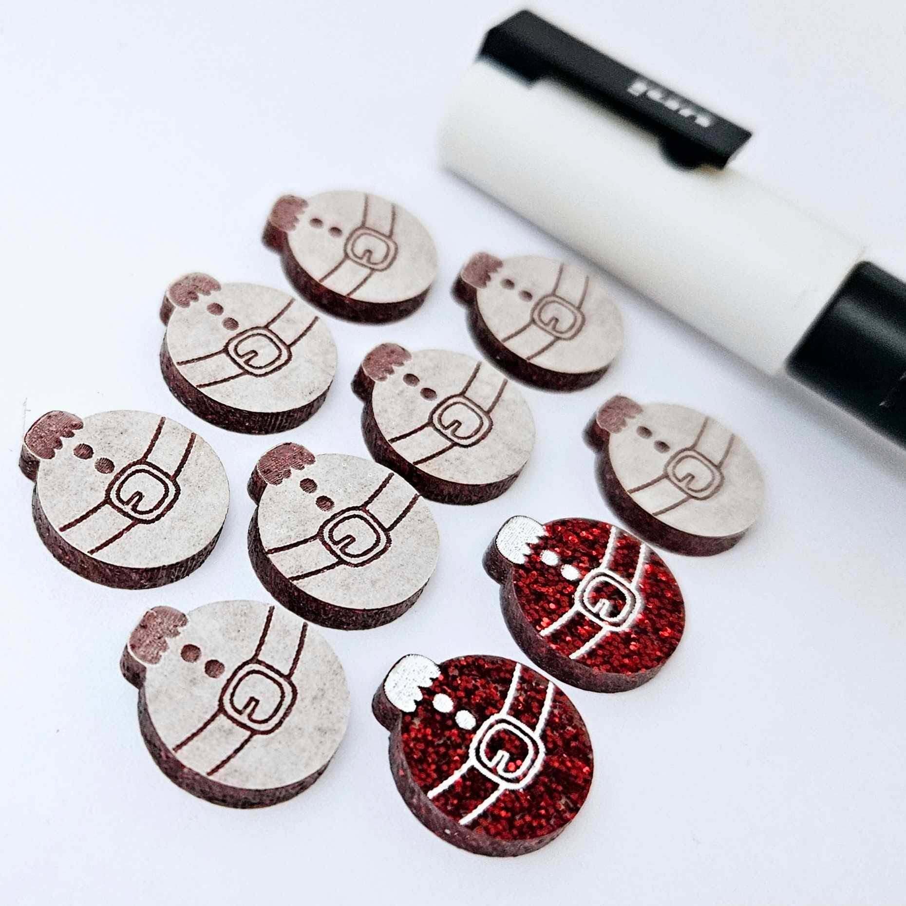 One pair of red glitter Santa bauble stud blanks paint filled white, surrounded by multiple unpainted blanks with protective paper attached, pictured with a Posca paint pen