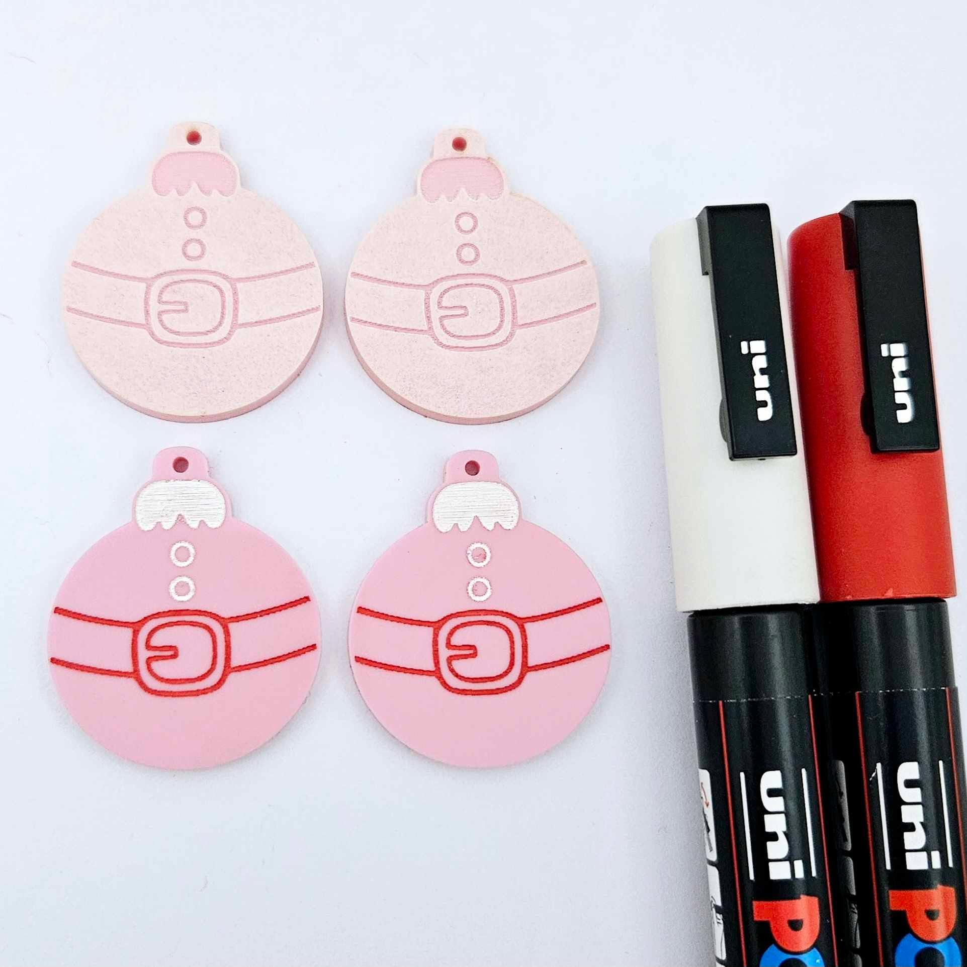 One pair of pastel pink Santa bauble dangle blanks paint filled in white and red, alongside one unpainted pair with protective paper attached, pictured with Posca paint pens