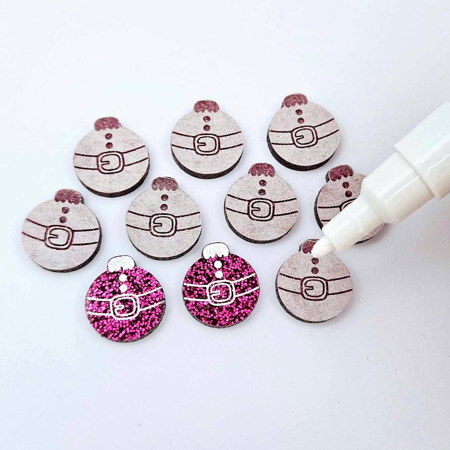 One pair of hot pink glitter Santa bauble stud blanks paint filled white alongside multiple unpainted blanks with protective paper attached, with an uncapped Posca paint pen positioned ready to paint fill the engraved details