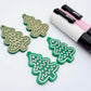 One pair of solid green cookie Christmas tree dangle earring blanks paint filled in light pink and white, alongside one unpainted pair with protective paper still attached, pictured with Posca paint pens