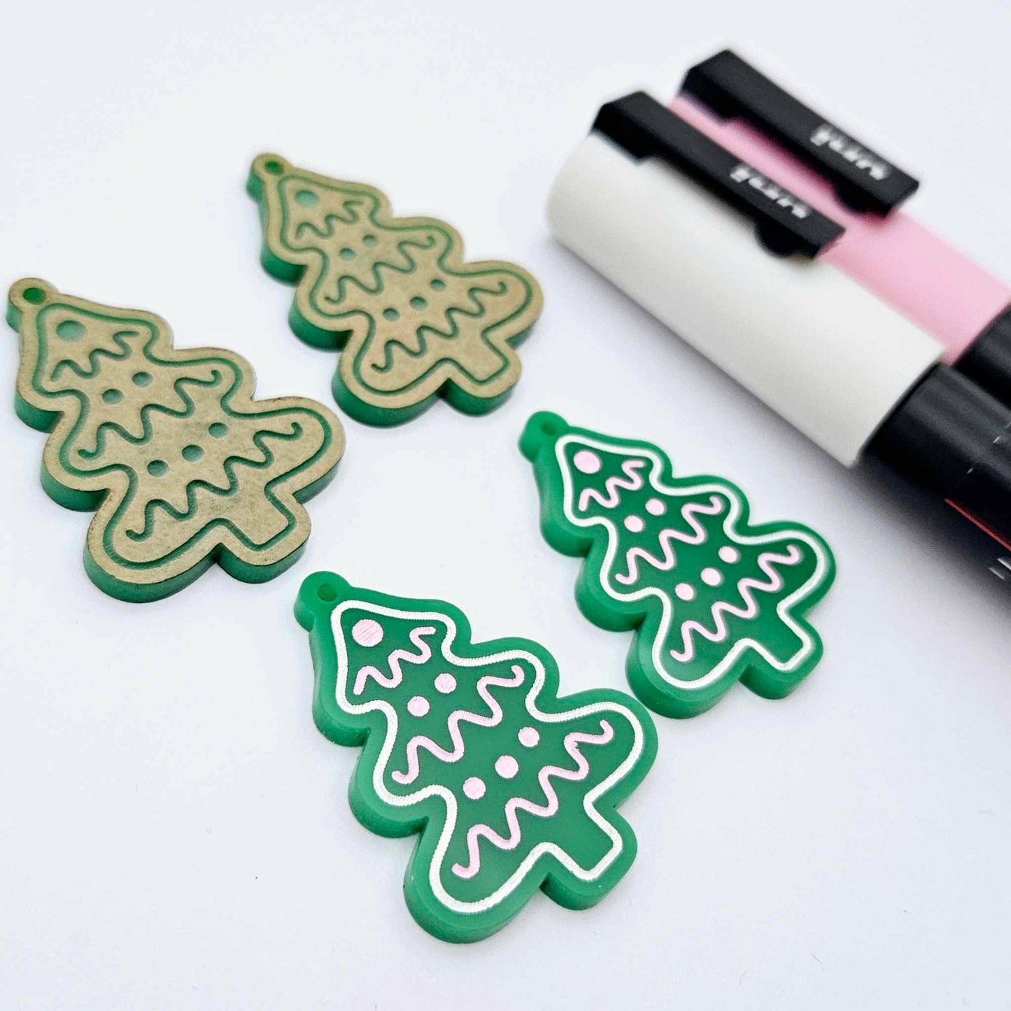 One pair of solid green cookie Christmas tree dangle earring blanks paint filled in light pink and white, alongside one unpainted pair with protective paper still attached, pictured with Posca paint pens