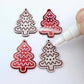 One pair of paint-filled deep red marble cookie Christmas tree dangle blanks beside one unpainted pair with protective paper attached, with an uncapped Posca paint pen positioned ready to paint fill the engraved details