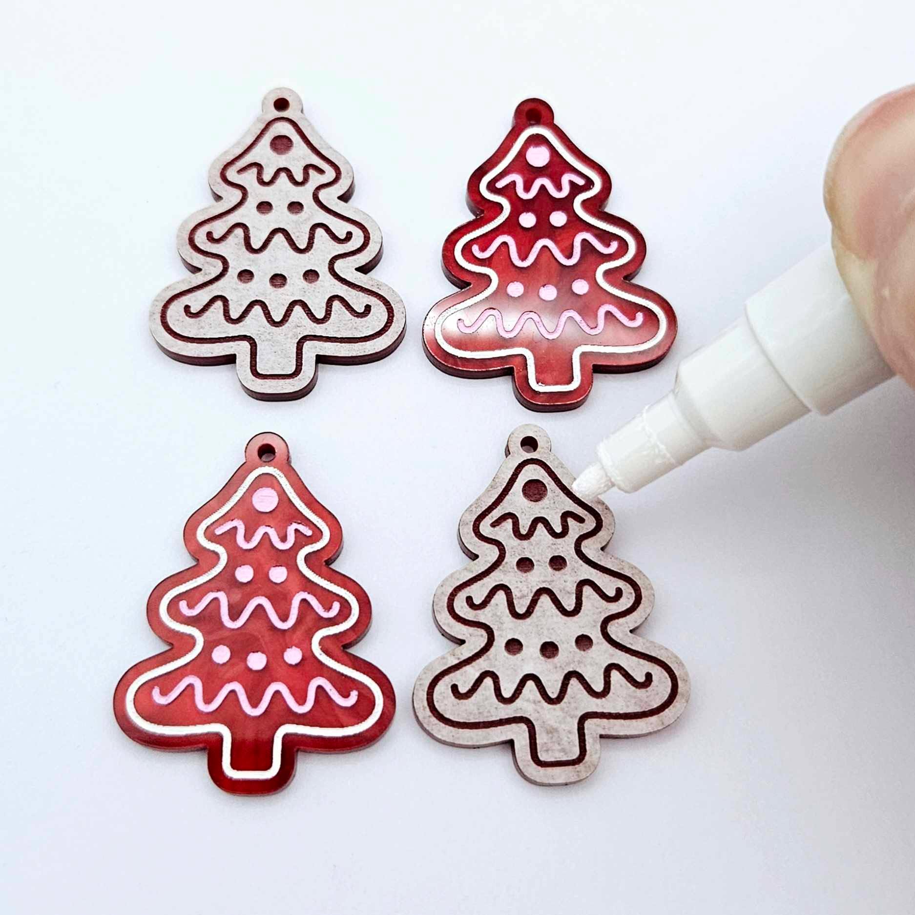 One pair of paint-filled deep red marble cookie Christmas tree dangle blanks beside one unpainted pair with protective paper attached, with an uncapped Posca paint pen positioned ready to paint fill the engraved details