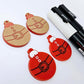 One pair of solid red Santa bauble dangle earring blanks paint filled in black and white, alongside one unpainted pair with protective paper attached, pictured with Posca paint pens