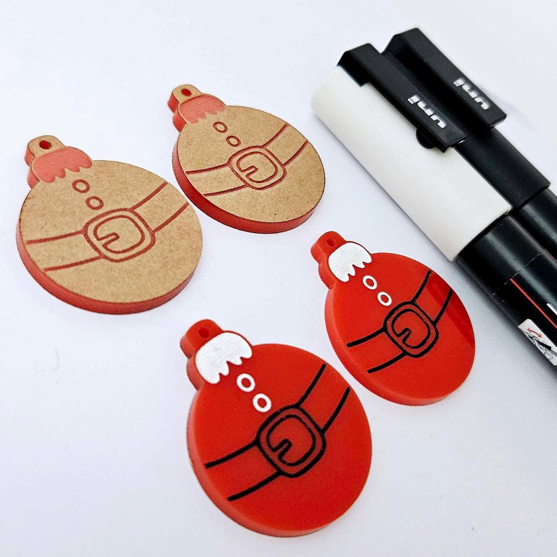 One pair of solid red Santa bauble dangle earring blanks paint filled in black and white, alongside one unpainted pair with protective paper attached, pictured with Posca paint pens