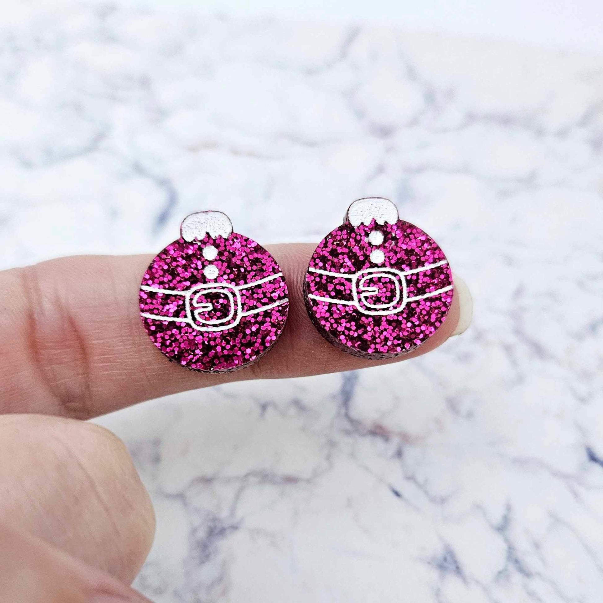One pair of hot pink glitter Santa bauble stud earring blanks with engraved Santa belt and button details paint filled white, shown on a finger for size reference