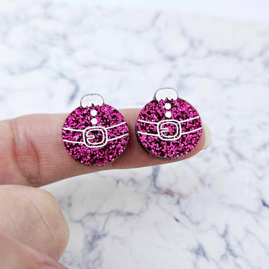 One pair of hot pink glitter Santa bauble stud earring blanks with engraved Santa belt and button details paint filled white, shown on a finger for size reference