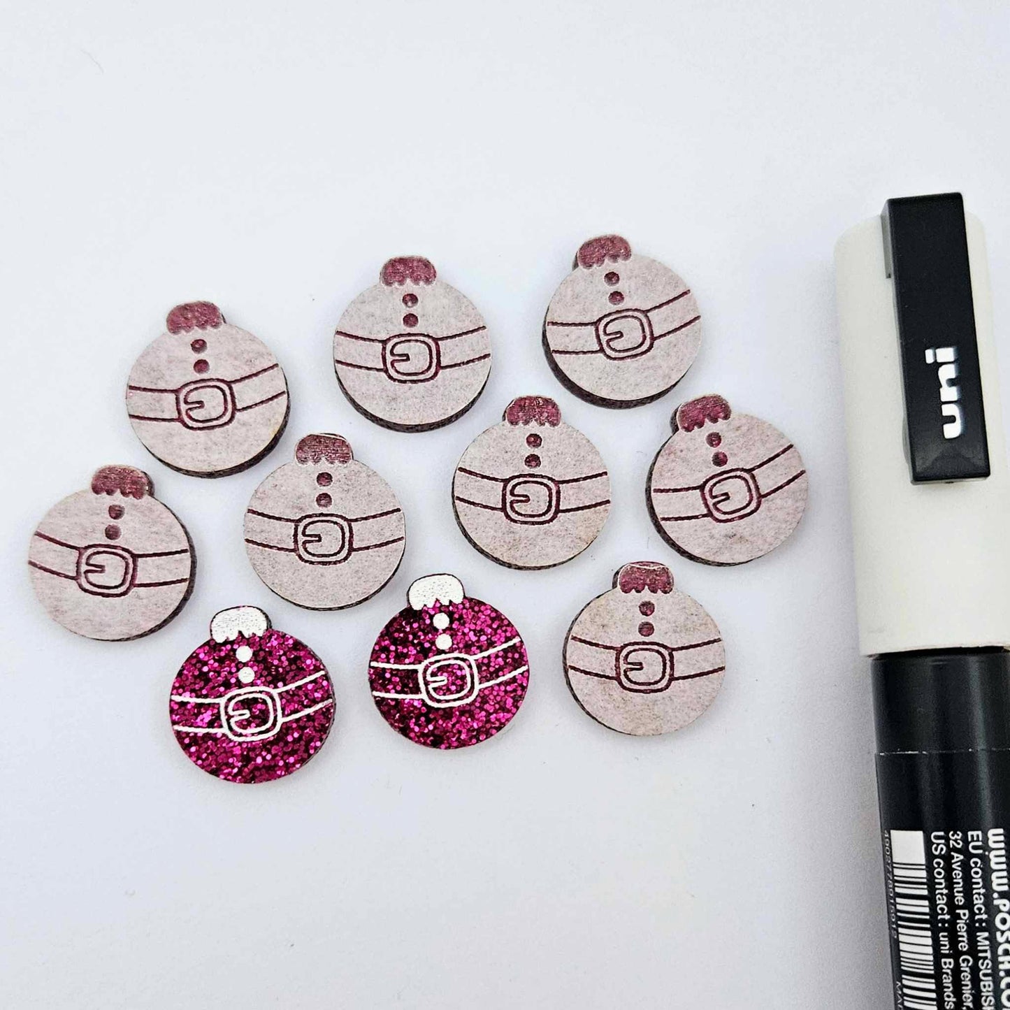 One pair of hot pink glitter Santa bauble stud blanks paint filled white, surrounded by multiple unpainted blanks with protective paper attached, pictured with a Posca paint pen