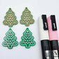 One pair of solid green cookie Christmas tree dangle earring blanks paint filled in light pink and white, alongside one unpainted pair with protective paper still attached, pictured with Posca paint pens
