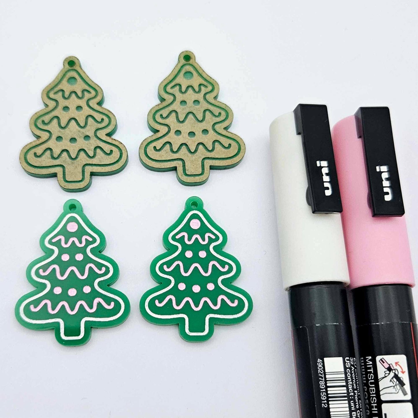 One pair of solid green cookie Christmas tree dangle earring blanks paint filled in light pink and white, alongside one unpainted pair with protective paper still attached, pictured with Posca paint pens