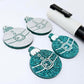 One pair of teal glitter Santa bauble dangle blanks paint filled white, alongside one unpainted pair with protective paper attached, pictured with  a Posca paint pen
