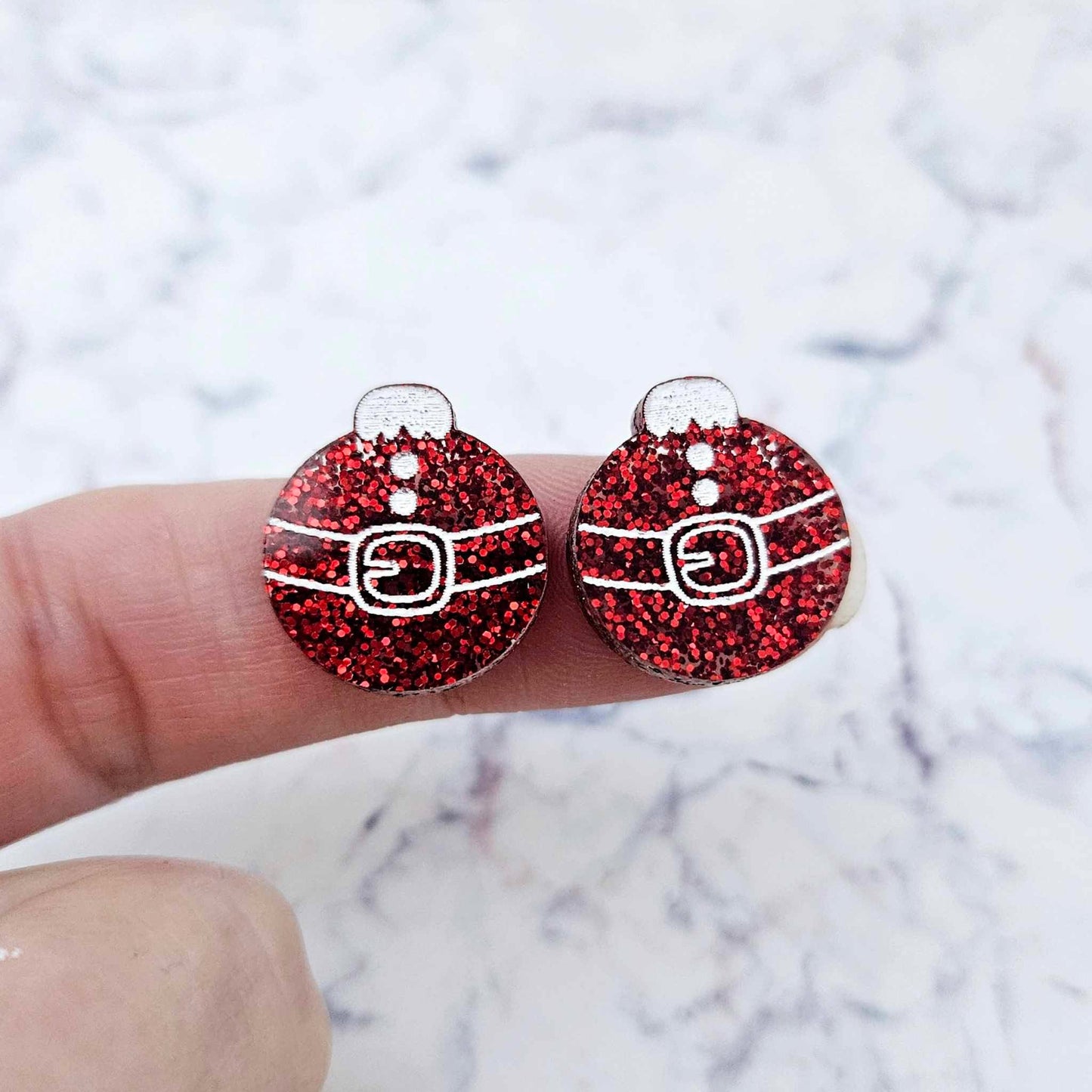 One pair of red glitter Santa bauble stud earrings blanks with engraved Santa belt and button details paint filled white, shown on a finger for size reference