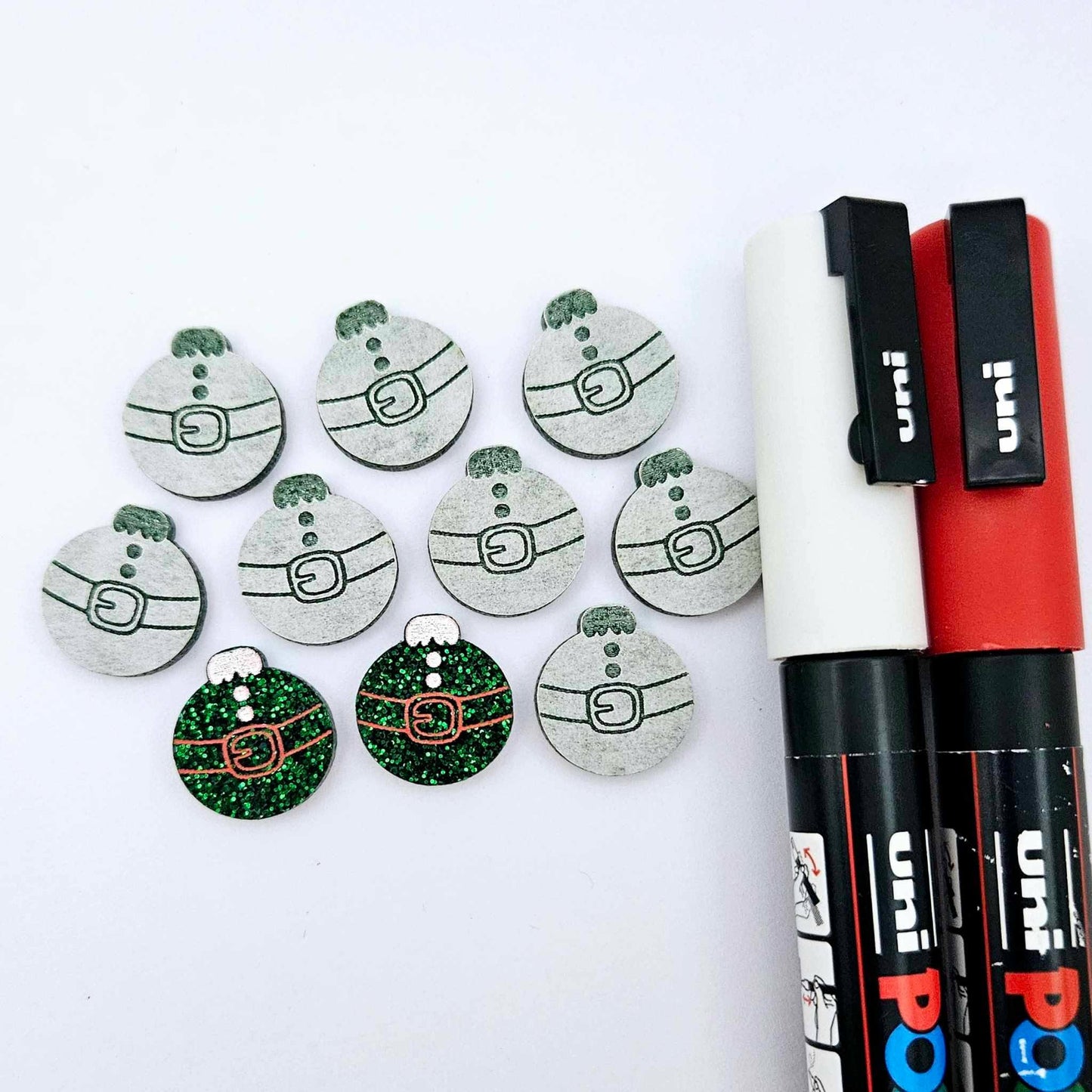 One pair of green glitter Santa bauble stud blanks paint filled red and white, surrounded by multiple unpainted blanks with protective paper attached, pictured with Posca paint pens