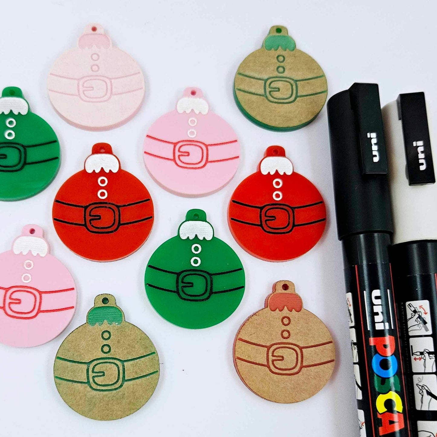 One paint-filled pair each of solid red, solid green and pastel pink Santa bauble dangle blanks, with additional unpainted blanks still covered with protective paper scattered around, showing the paint-fill possibilities.