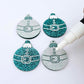 One pair of paint-filled teal glitter Santa bauble dangle blanks with one unpainted pair with protective paper attached, with an uncapped Posca paint pen positioned ready to paint fill the engraved Santa belt details