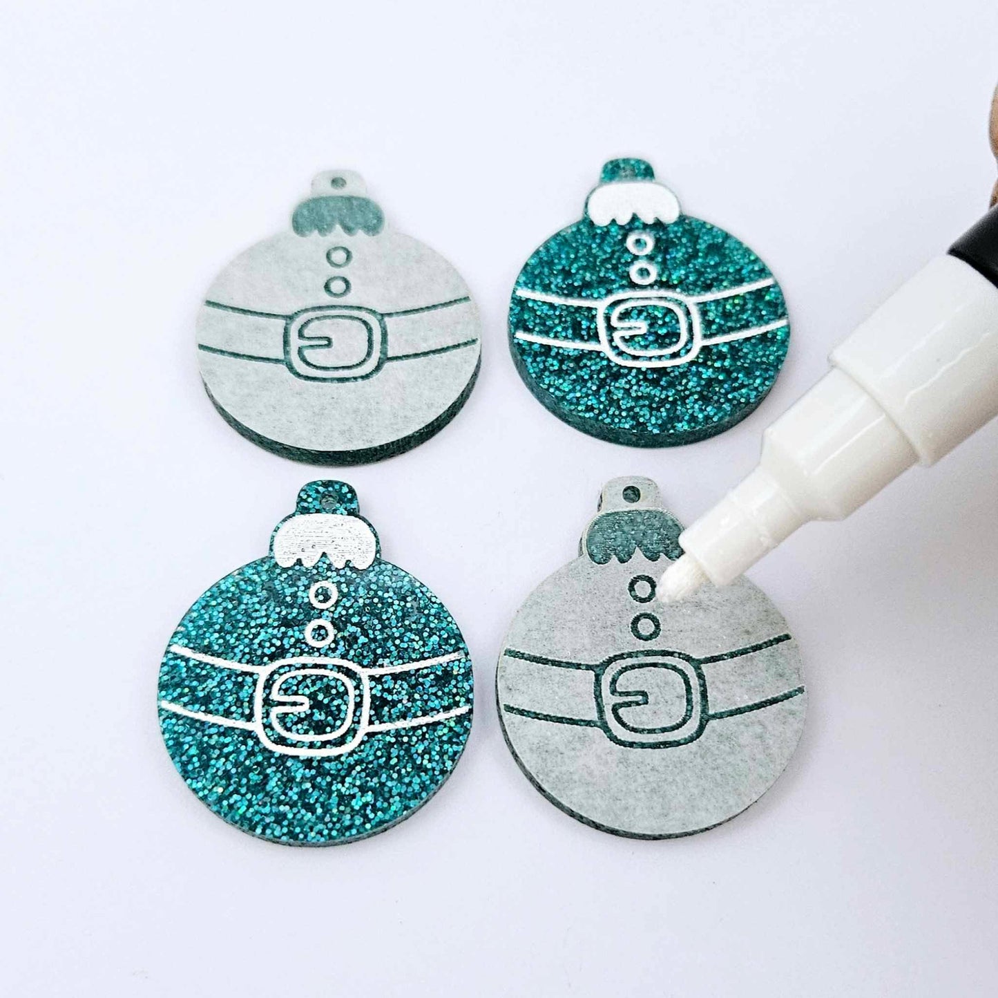 One pair of paint-filled teal glitter Santa bauble dangle blanks with one unpainted pair with protective paper attached, with an uncapped Posca paint pen positioned ready to paint fill the engraved Santa belt details