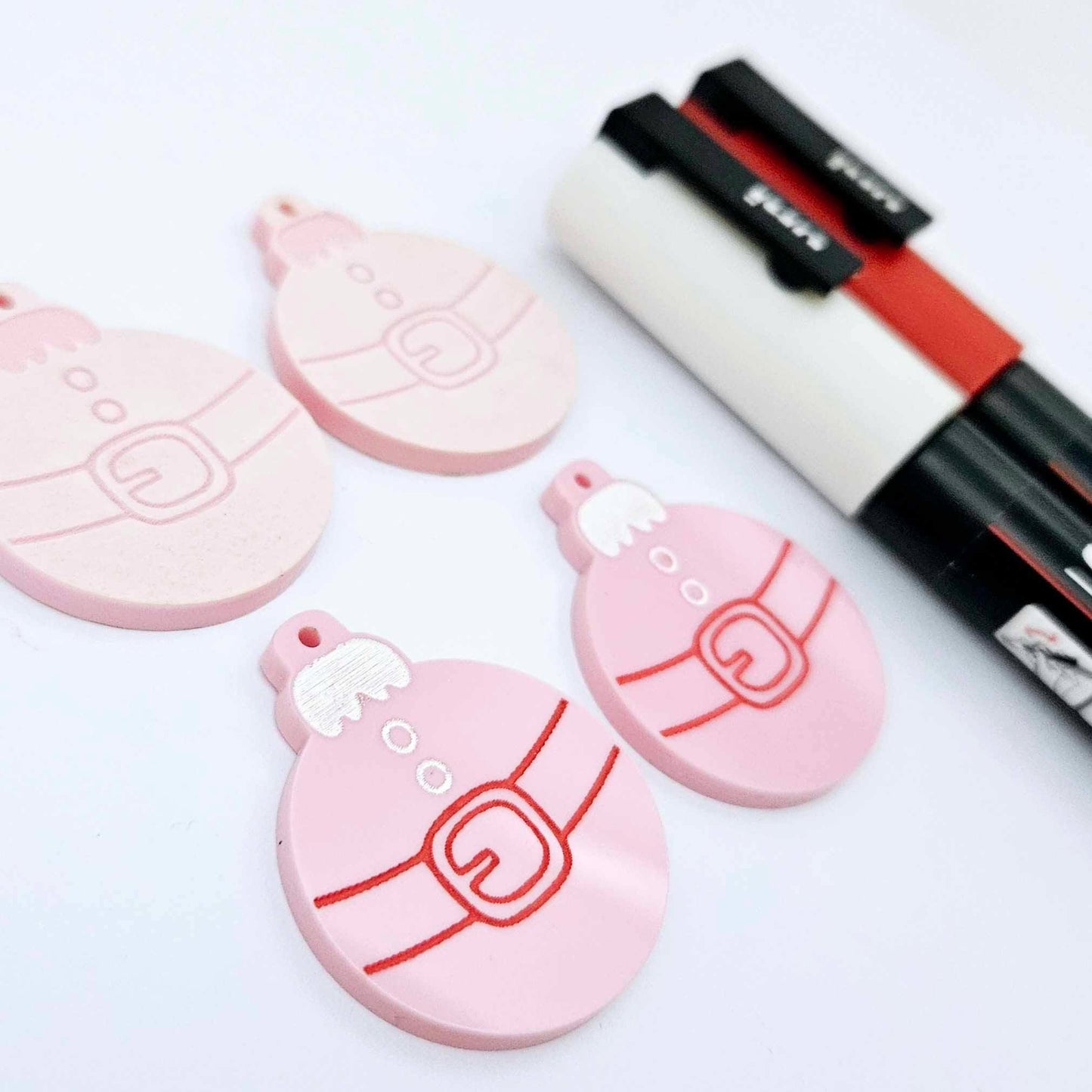 One pair of pastel pink Santa bauble dangle blanks paint filled in white and red, alongside one unpainted pair with protective paper attached, pictured with Posca paint pens