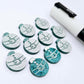 One pair of teal glitter Santa bauble stud blanks paint filled white, surrounded by multiple unpainted blanks with protective paper attached, pictured with a Posca paint pen