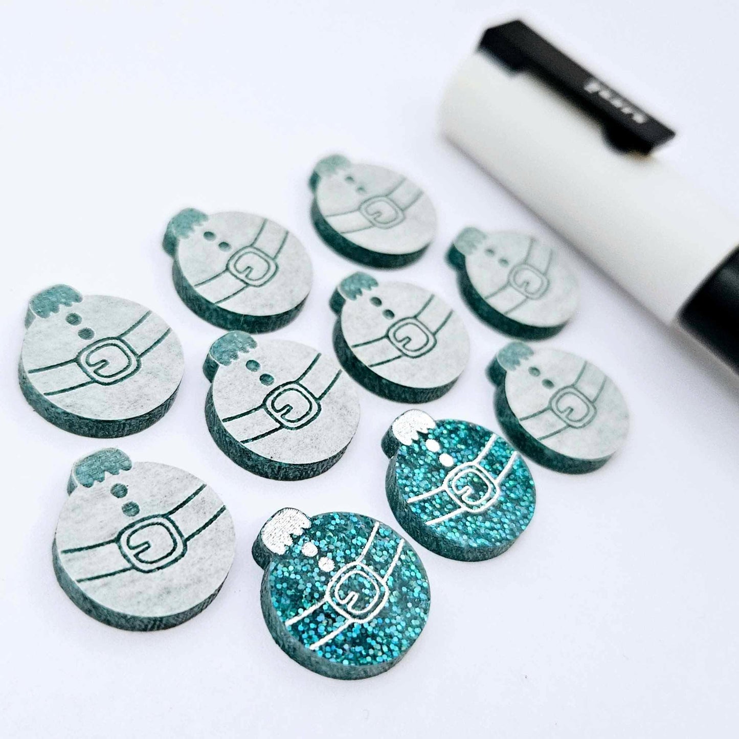 One pair of teal glitter Santa bauble stud blanks paint filled white, surrounded by multiple unpainted blanks with protective paper attached, pictured with a Posca paint pen