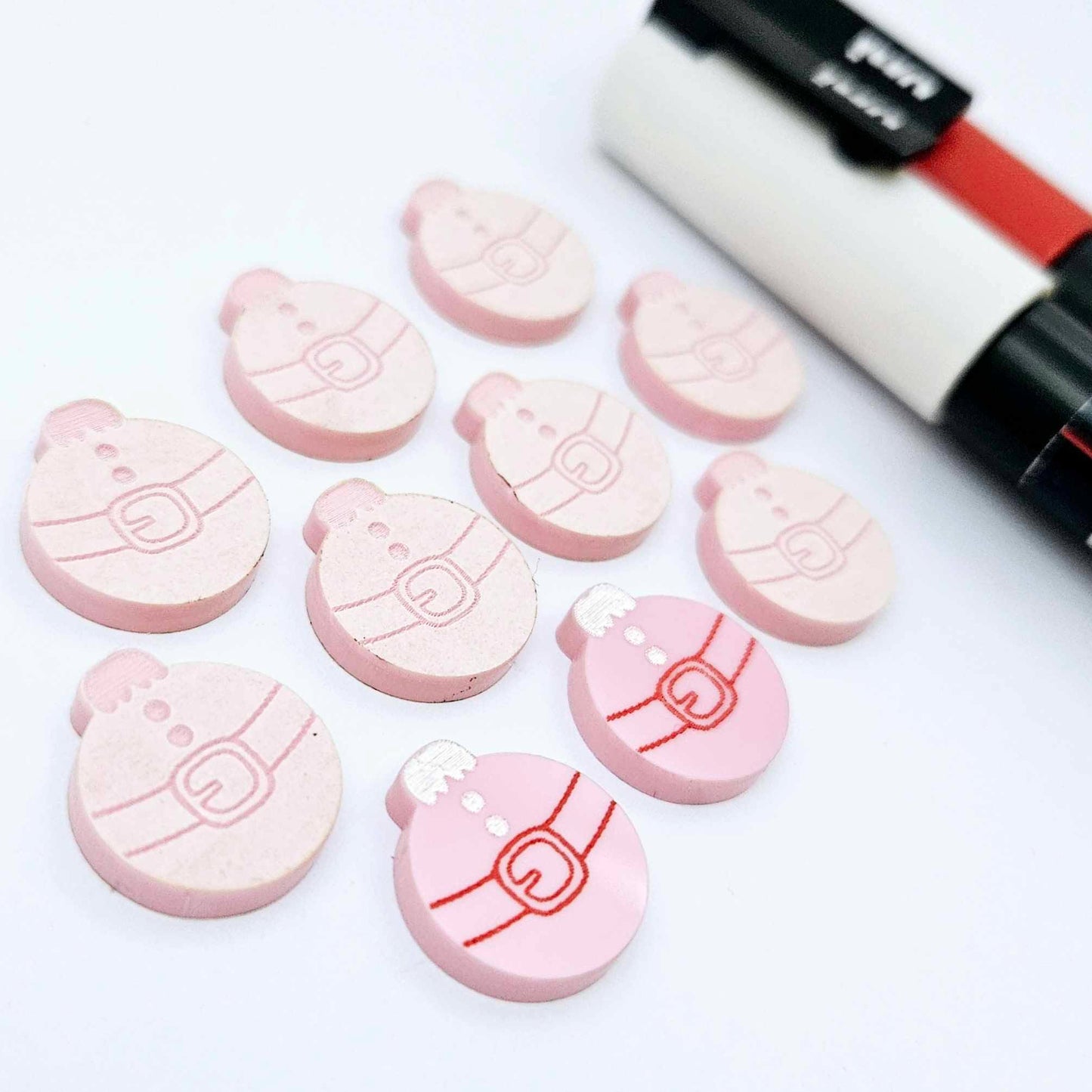 One pair of pastel pink Santa bauble stud blanks paint filled red and white, surrounded by multiple unpainted blanks with protective paper attached, pictured with Posca paint pens