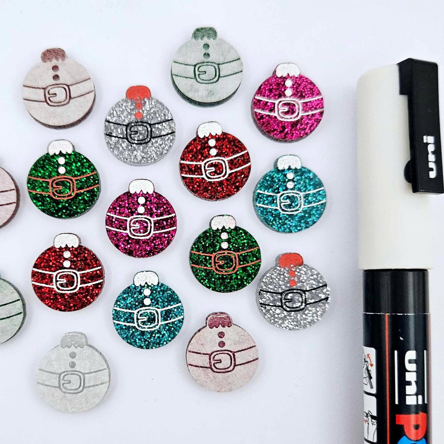 One paint-filled pair each of red glitter, hot pink glitter, teal glitter, green glitter and silver glitter Santa bauble stud blanks, with additional unpainted blanks still covered with protective paper scattered around, showing the paint-fill possibilities