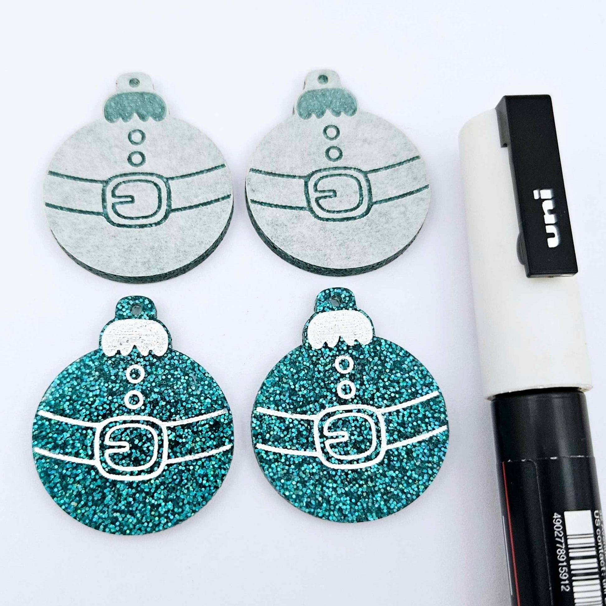 One pair of teal glitter Santa bauble dangle blanks paint filled white, alongside one unpainted pair with protective paper attached, pictured with  a Posca paint pen
