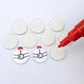 One pair of white marble Santa bauble stud blanks paint filled red and black alongside multiple unpainted blanks with protective paper attached, with an uncapped Posca paint pen positioned ready to paint fill the engraved details