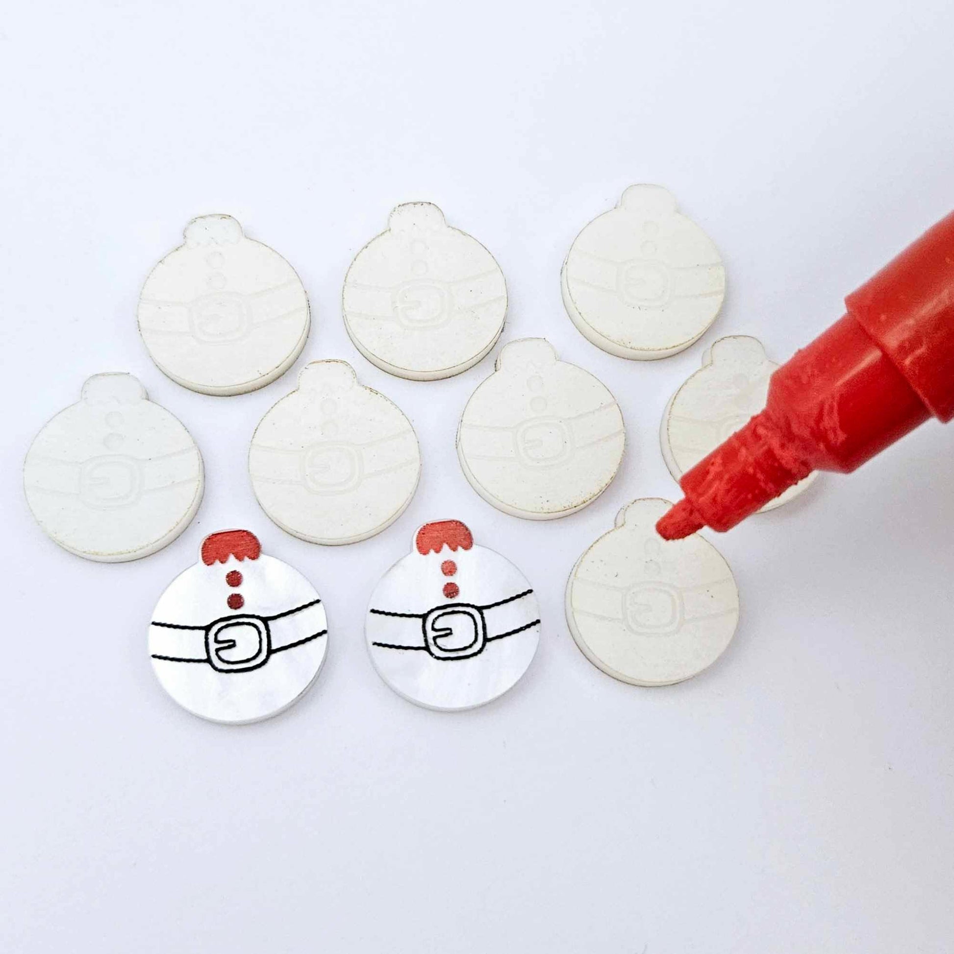 One pair of white marble Santa bauble stud blanks paint filled red and black alongside multiple unpainted blanks with protective paper attached, with an uncapped Posca paint pen positioned ready to paint fill the engraved details