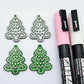 One pair of green marble cookie Christmas tree dangle blanks paint filled in light pink and white, alongside one unpainted pair with protective paper attached, pictured with Posca paint pens