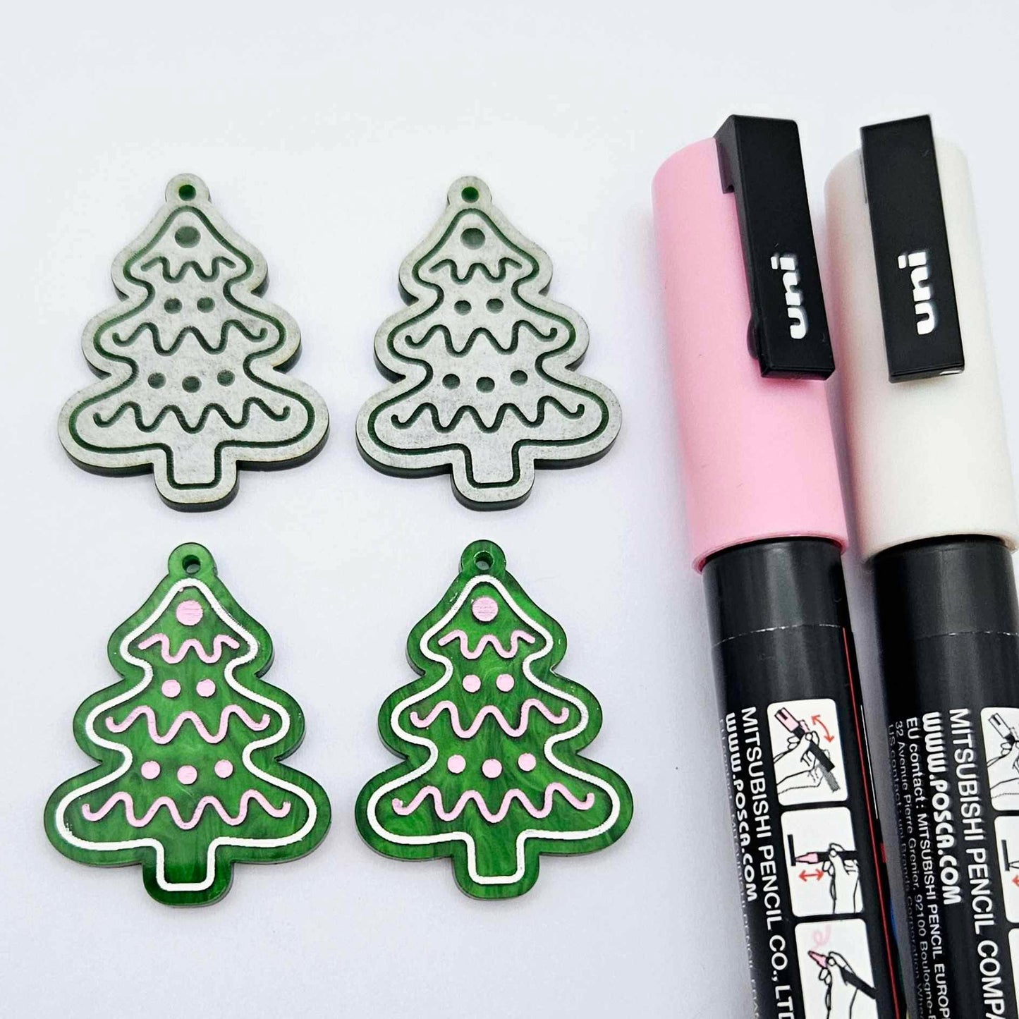 One pair of green marble cookie Christmas tree dangle blanks paint filled in light pink and white, alongside one unpainted pair with protective paper attached, pictured with Posca paint pens