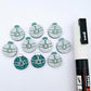 One pair of teal glitter Santa bauble stud blanks paint filled white, surrounded by multiple unpainted blanks with protective paper attached, pictured with a Posca paint pen