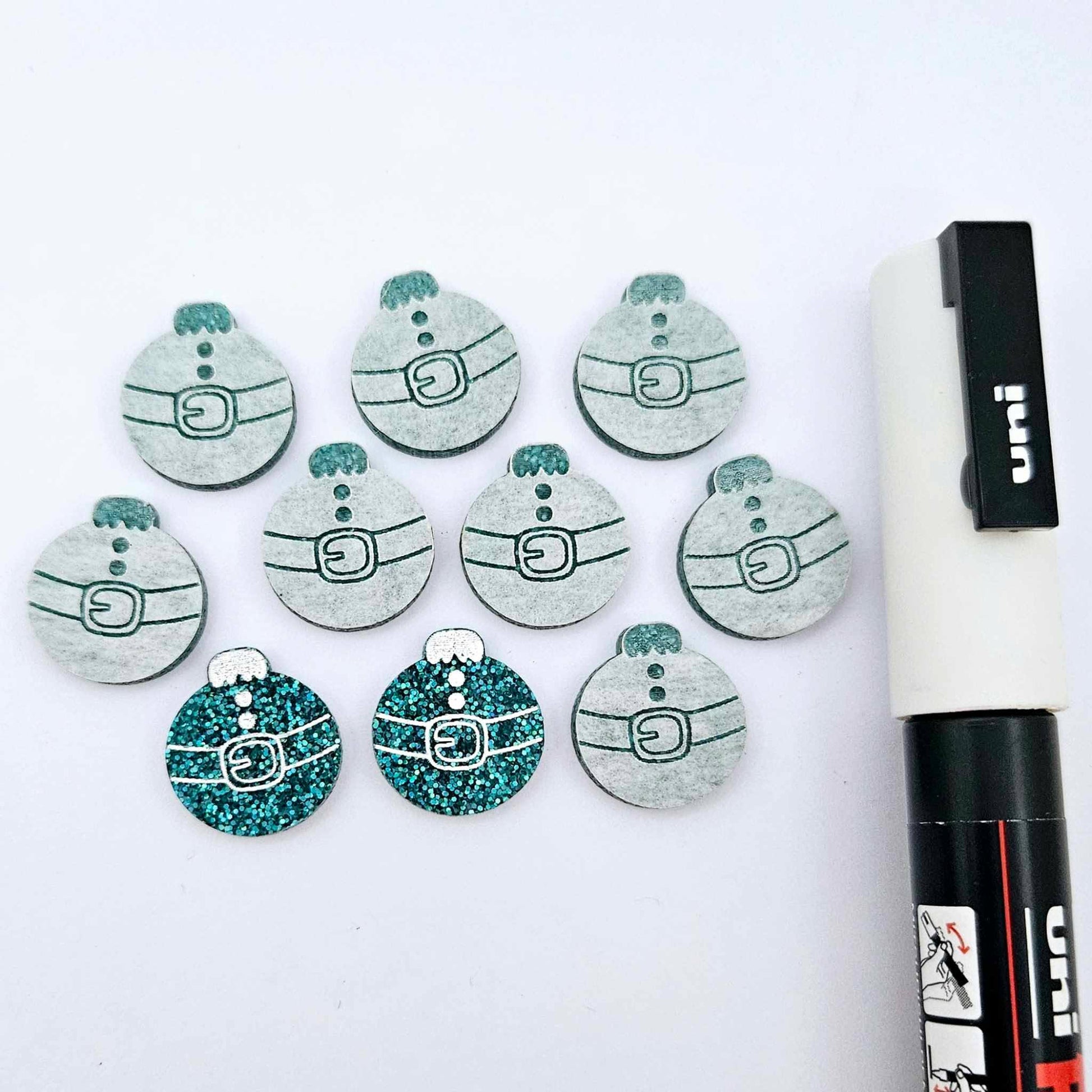 One pair of teal glitter Santa bauble stud blanks paint filled white, surrounded by multiple unpainted blanks with protective paper attached, pictured with a Posca paint pen