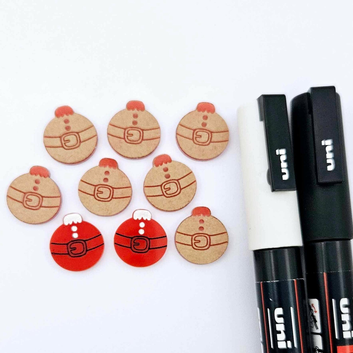 One pair of solid red Santa bauble stud blanks paint filled black and white, surrounded by multiple unpainted blanks with protective paper attached, pictured with Posca paint pens
