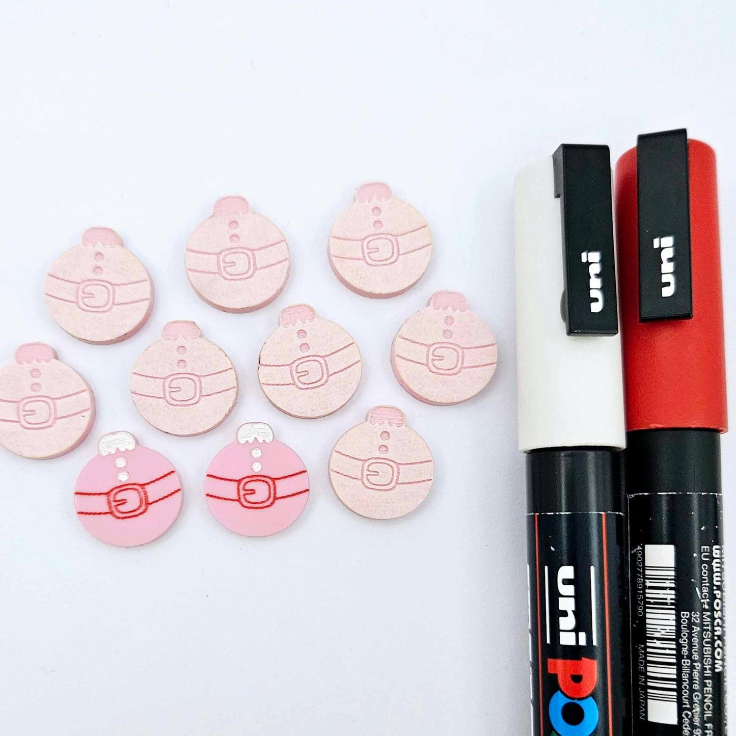 One pair of pastel pink Santa bauble stud blanks paint filled red and white, surrounded by multiple unpainted blanks with protective paper attached, pictured with Posca paint pens