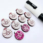One pair of hot pink glitter Santa bauble stud blanks paint filled white, surrounded by multiple unpainted blanks with protective paper attached, pictured with a Posca paint pen