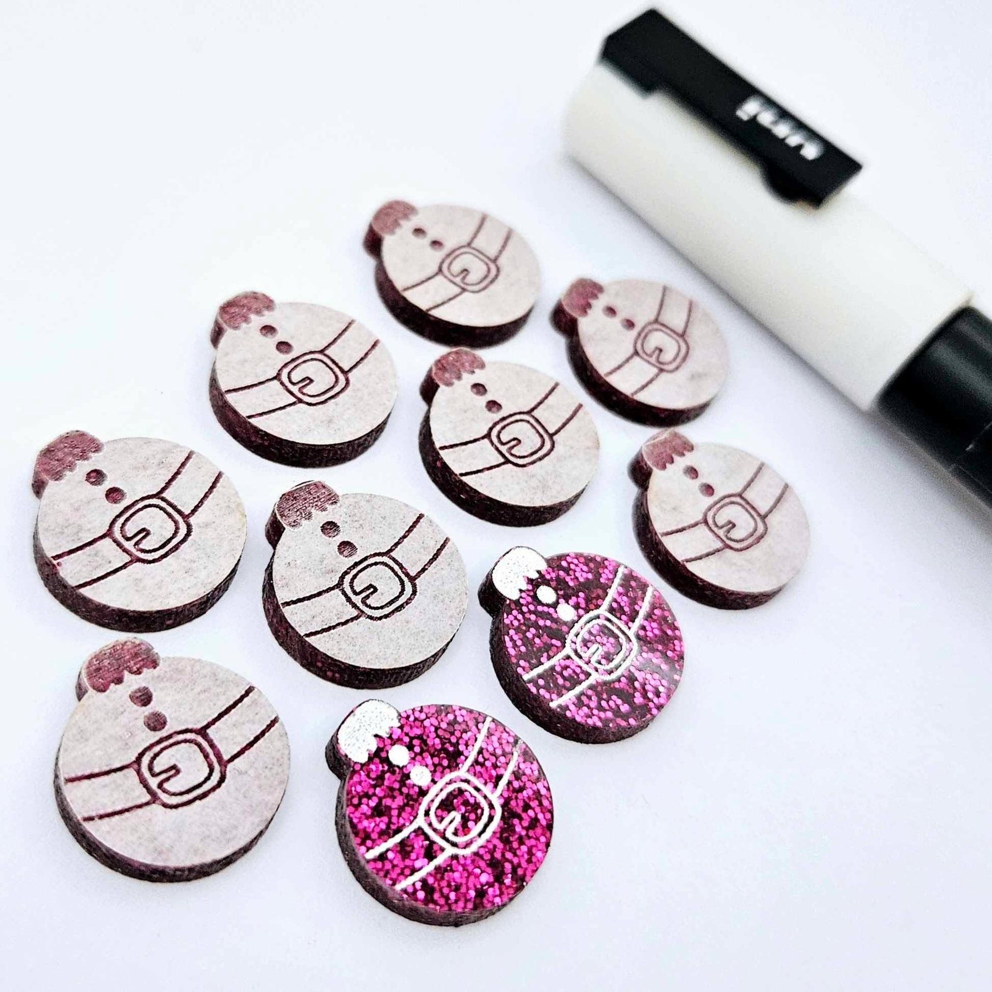 One pair of hot pink glitter Santa bauble stud blanks paint filled white, surrounded by multiple unpainted blanks with protective paper attached, pictured with a Posca paint pen