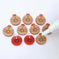One pair of solid red Santa bauble stud blanks paint filled black and white alongside multiple unpainted blanks with protective paper attached, with an uncapped Posca paint pen positioned ready to paint fill the engraved details