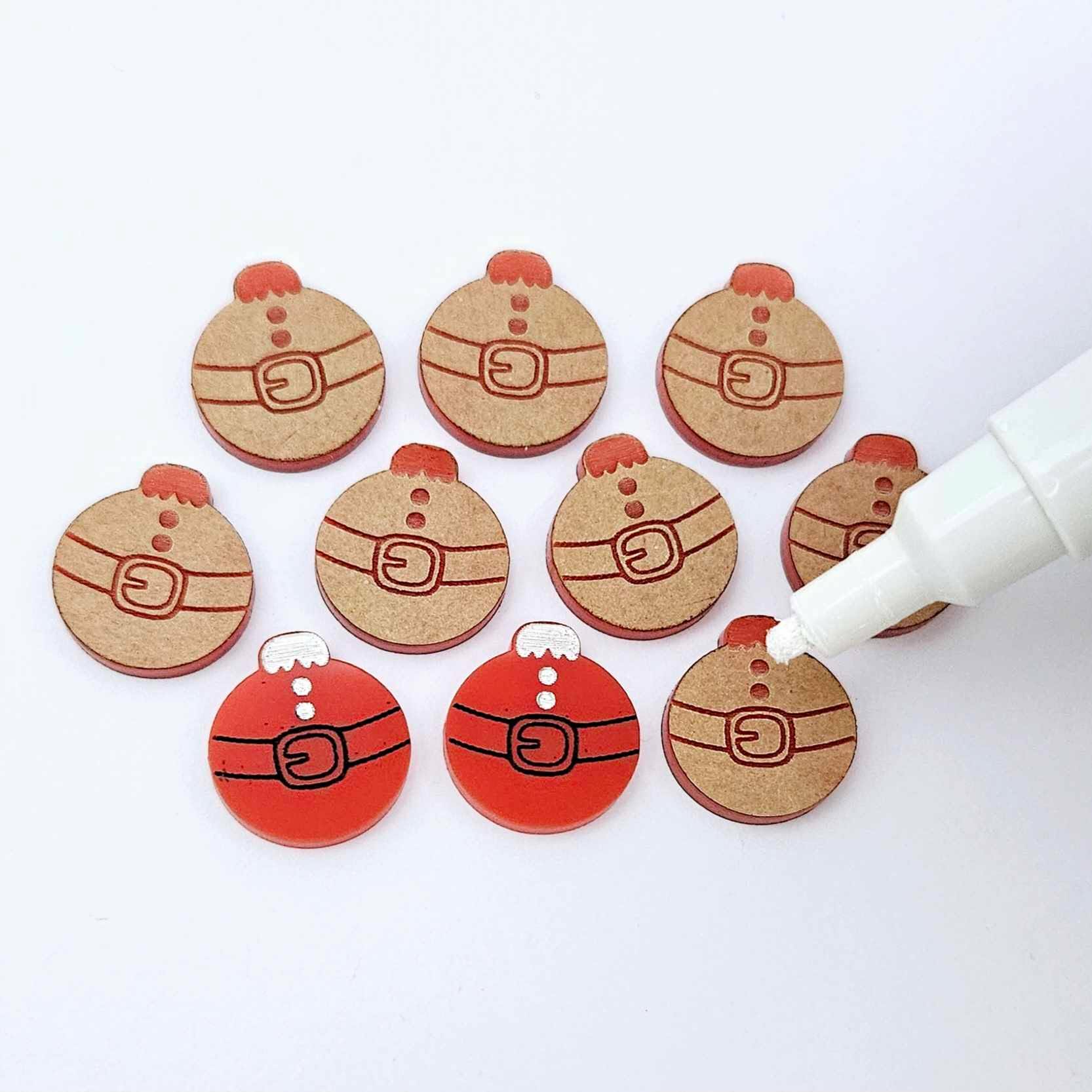 One pair of solid red Santa bauble stud blanks paint filled black and white alongside multiple unpainted blanks with protective paper attached, with an uncapped Posca paint pen positioned ready to paint fill the engraved details
