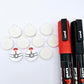 One pair of white marble Santa bauble stud blanks paint filled red and black, surrounded by multiple unpainted blanks with protective paper attached, pictured with Posca paint pens