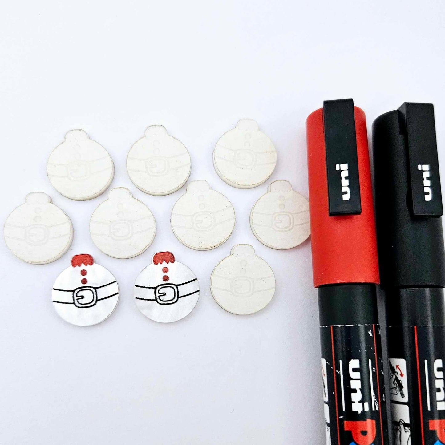 One pair of white marble Santa bauble stud blanks paint filled red and black, surrounded by multiple unpainted blanks with protective paper attached, pictured with Posca paint pens