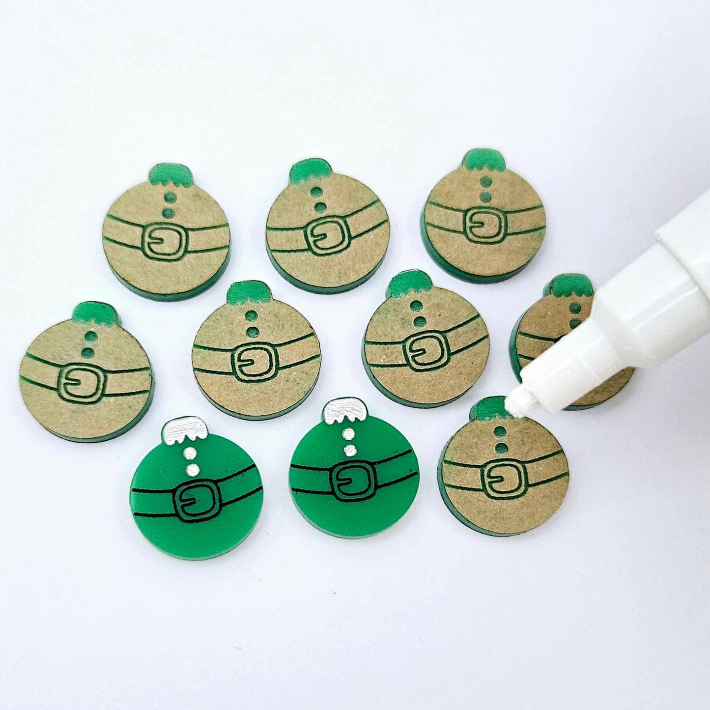 One pair of solid green Santa bauble stud blanks paint filled black and white alongside multiple unpainted blanks with protective paper attached, with an uncapped Posca paint pen positioned ready to paint fill the engraved details
