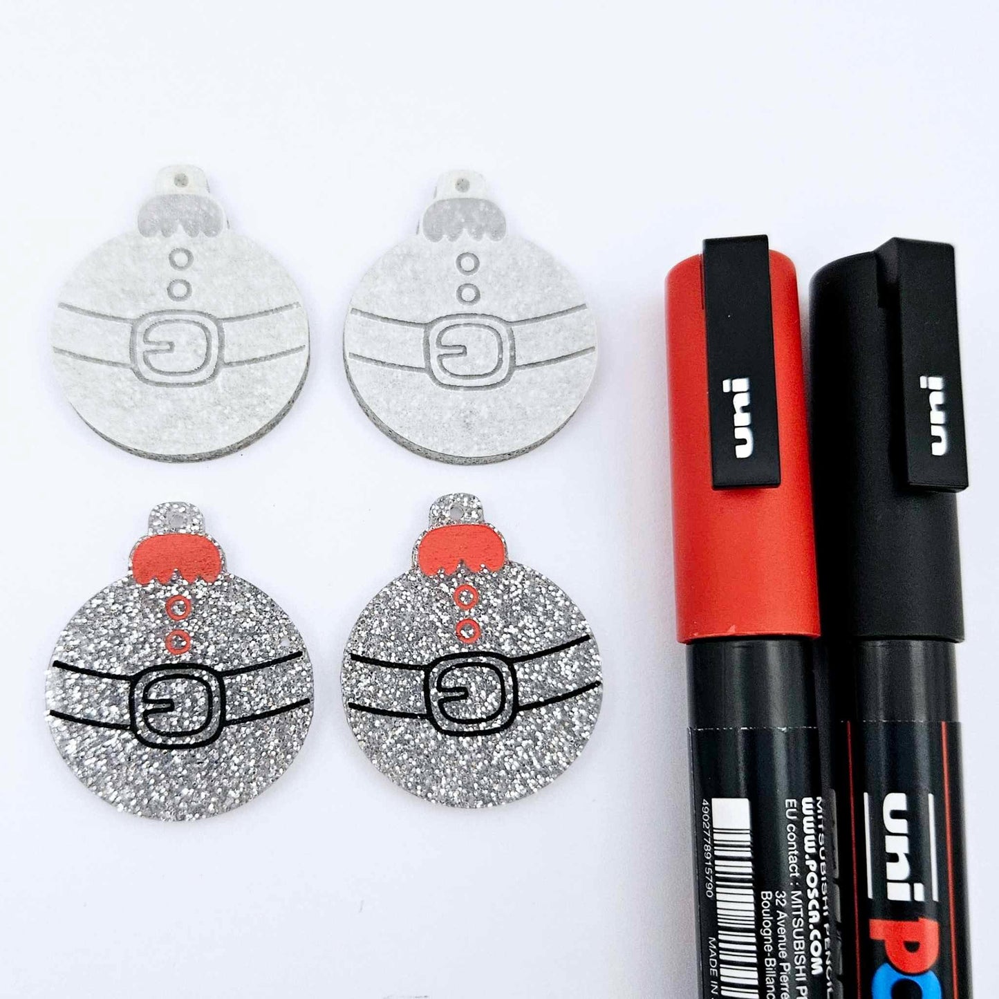 One pair of silver glitter Santa bauble dangle blanks paint filled red and black, alongside one unpainted pair with protective paper attached, pictured with Posca paint pens