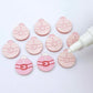 One pair of pastel pink Santa bauble stud blanks paint filled red and white alongside multiple unpainted blanks with protective paper attached, with an uncapped Posca paint pen positioned ready to paint fill the engraved details