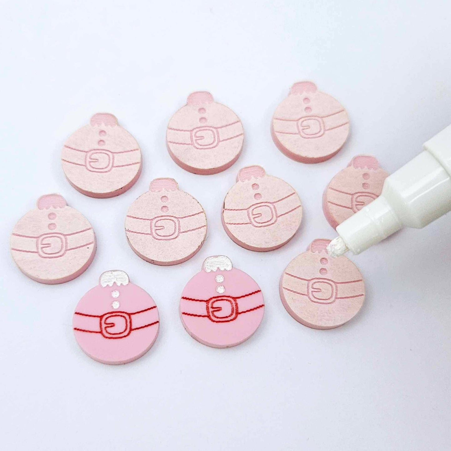 One pair of pastel pink Santa bauble stud blanks paint filled red and white alongside multiple unpainted blanks with protective paper attached, with an uncapped Posca paint pen positioned ready to paint fill the engraved details