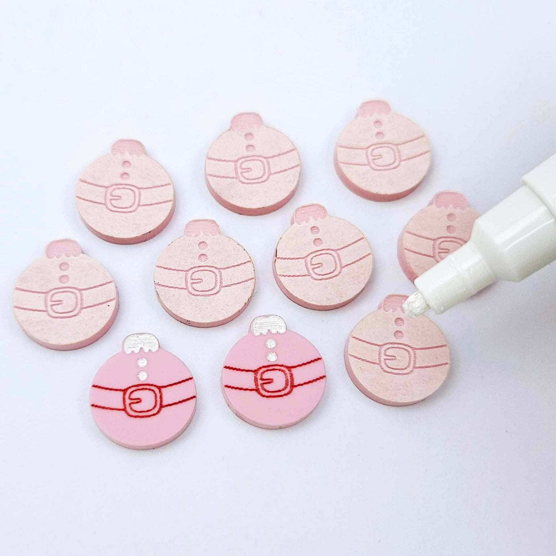 One pair of pastel pink Santa bauble stud blanks paint filled red and white alongside multiple unpainted blanks with protective paper attached, with an uncapped Posca paint pen positioned ready to paint fill the engraved details