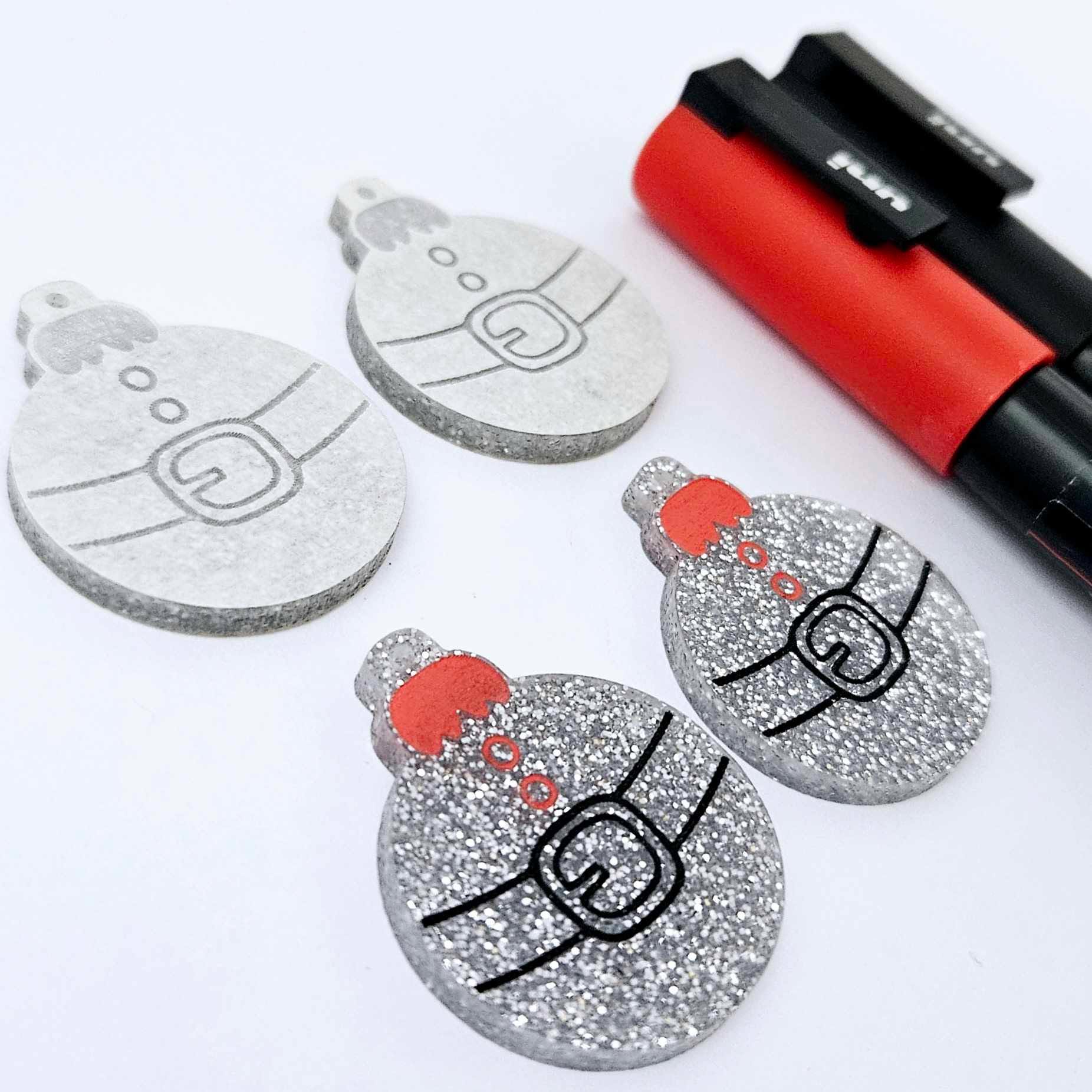 One pair of silver glitter Santa bauble dangle blanks paint filled red and black, alongside one unpainted pair with protective paper attached, pictured with Posca paint pens