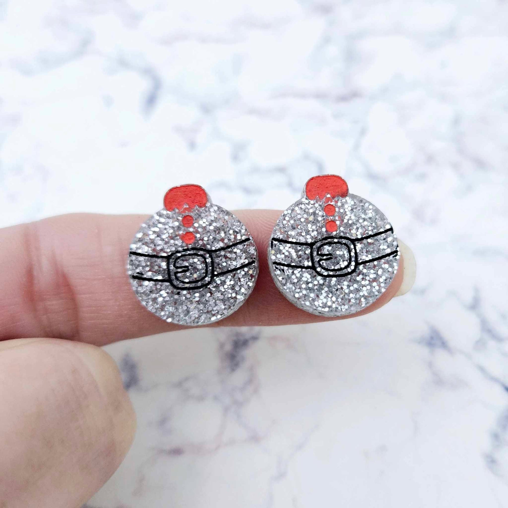 One pair of silver glitter Santa bauble stud earring blanks with engraved Santa belt and button details paint filled black and red, shown on a finger for size reference