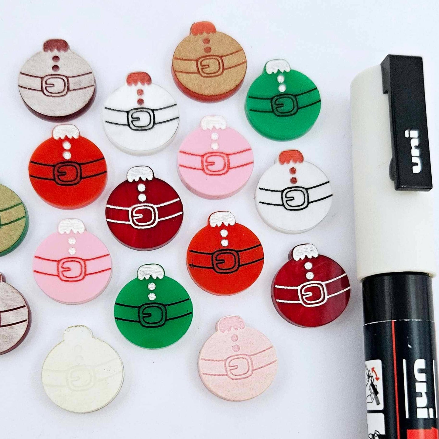One paint-filled pair each of solid red, solid green, pastel pink, white marble and red marble Santa bauble stud blanks, with additional unpainted blanks still covered with protective paper scattered around, showing the paint-fill possibilities